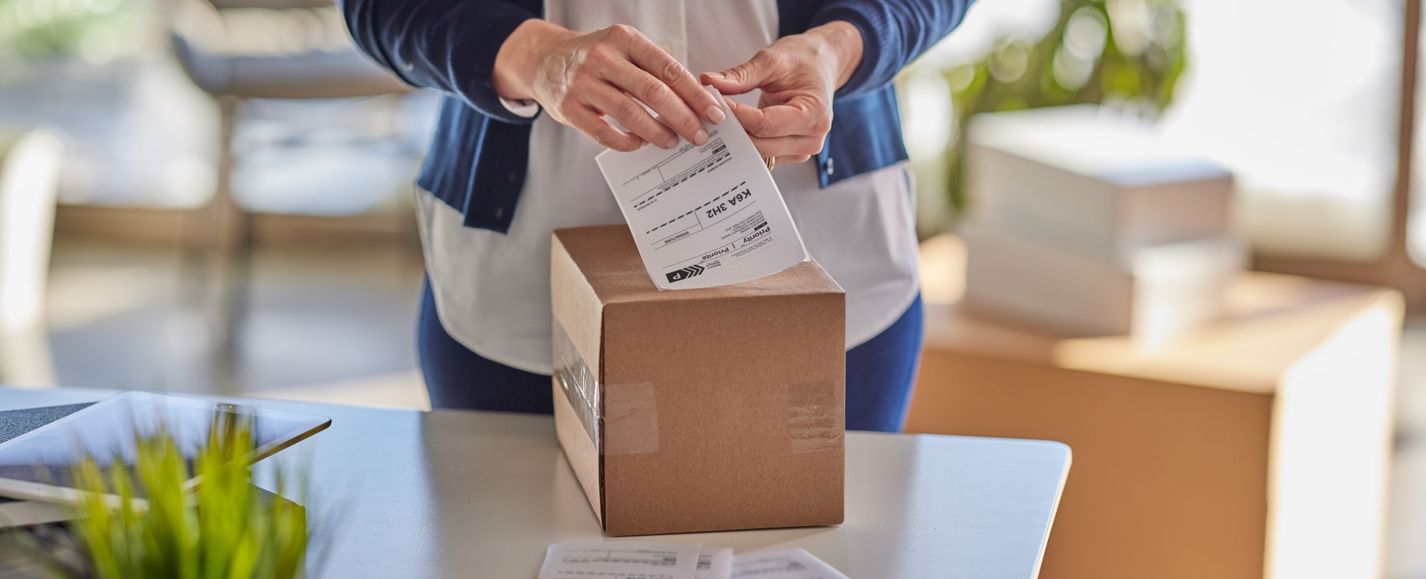 A woman stands in front of a table and applies a shipping label to a cardboard box.