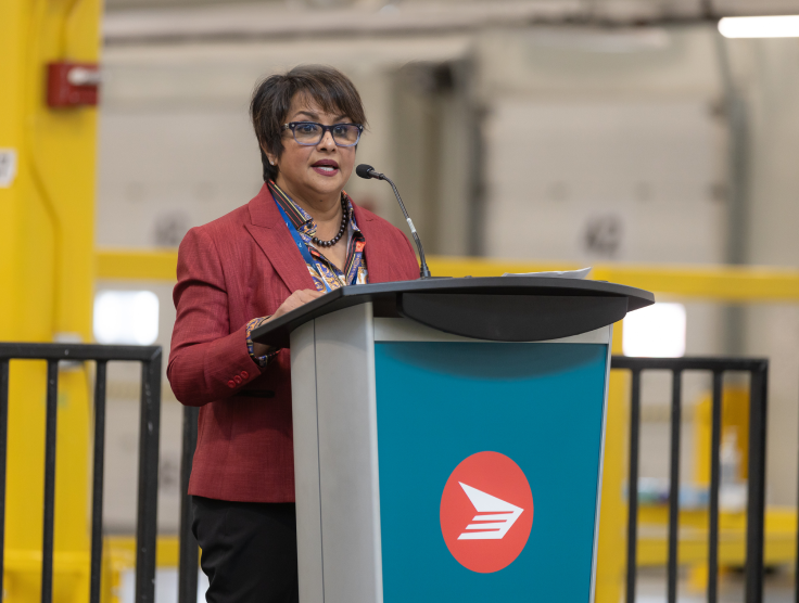 Canada Post's Chair of the Board of Directors, Suromitra Sanatani, stands and speaks at a podium inside the Albert Jackson Processing Centre.
