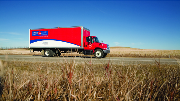 Red Canada Post truck on a road surrounded by a flat landscape of prairie grass and blue sky.