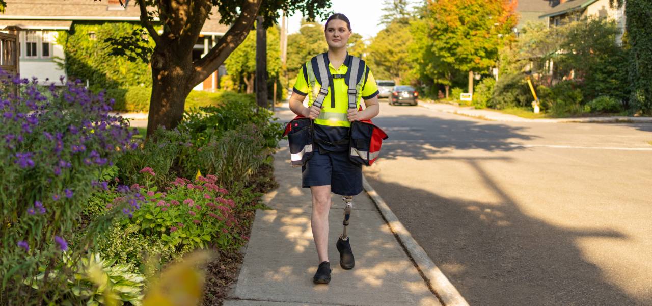 Une factrice de Postes Canada avec une jambe prothétique marche dans une rue résidentielle en portant des sacs de courrier. 